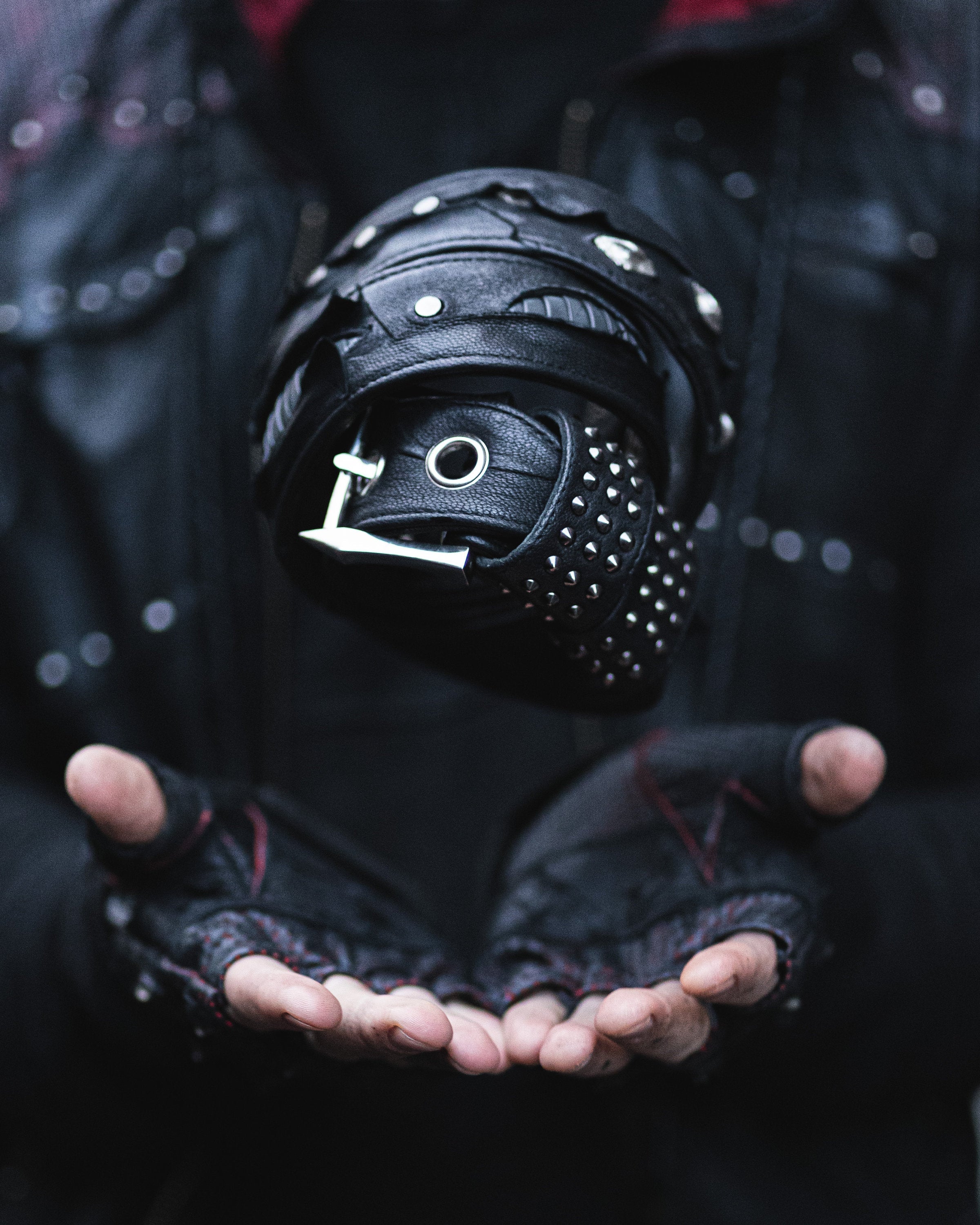 A person wearing dark clothing and fingerless gloves holds their hands out as a rolled-up Kaya Movement KRAV Wasteland Men's Black Studded Leather Belt with Silver Spine Studs hovers between their palms. The background is blurred, drawing focus to the belt and the person's hands.